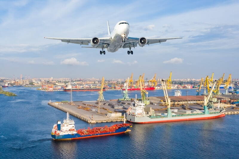 A charter airplane flying over a busy port with cranes and cargo ships below